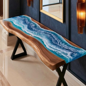 Könnte beinhalten: Ein Konsolentisch mit einer natürlichen Baumkanten-Holzplatte und einem zentralen Flussdesign aus blauem und weißem Harz, das Meereswellen nachahmt. Der Tisch wird von zwei schwarzen Metallbeinen mit geometrischem Rahmendesign getragen.