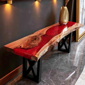 Puede incluir: Una mesa consola rectangular con un tablero de madera de borde natural y una incrustación central de resina roja translúcida. La mesa se apoya sobre dos patas de metal negro en forma de X y sostiene un jarrón dorado en su superficie.