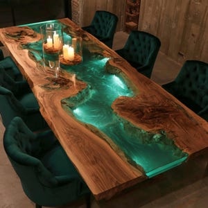 Puede incluir: Una mesa de comedor rectangular con un tablero de madera natural de borde vivo y una incrustación central tipo río de resina turquesa translúcida y brillante. La mesa está iluminada desde el interior y rodeada de sillas de terciopelo verde oscuro.