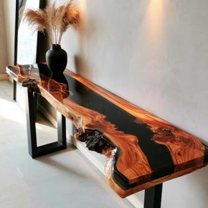 Peut inclure: Une longue table console composée d'une tranche de bois à bord naturel avec une incrustation centrale en résine noire. La table présente une finition brillante, des veines de bois apparentes et deux pieds rectangulaires en métal noir.