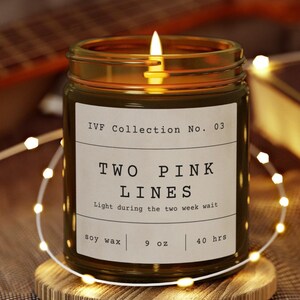 Op de afbeelding: Een brandende kaars in een glazen pot met de tekst "TWO PINK LINES" op een wit etiket. De kaars staat op een houten onderzetter, omringd door lichtslingers. Het etiket luidt ook "IVF Collection No. 03".