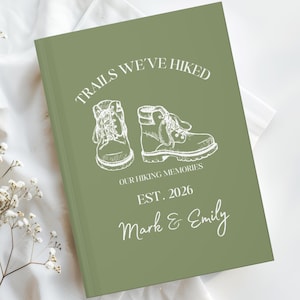 Könnte beinhalten: Ein olivgrünes Notizbuch mit dem Text "Trails We've Hiked" und einer Zeichnung von Wanderschuhen. Das Notizbuch enthält auch den Text "Our Hiking Memories" und "Est. 2026" mit den Namen "Mark & Emily" in Schreibschrift.