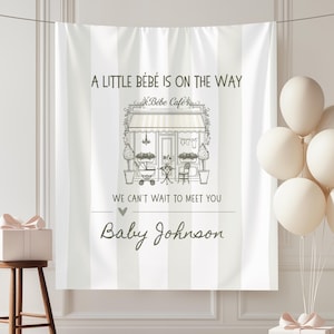 May include: A white and light gray vertical striped fabric backdrop featuring a graphic of a cafe storefront with the text A LITTLE BÉBÉ IS ON THE WAY, Bébé Café, WE CAN'T WAIT TO MEET YOU, and the personalized name Baby Johnson in dark gray.