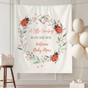 May include: A white backdrop with a floral wreath design featuring ladybugs and the text "A Little Lovebug Is On The Way Welcome Baby Piper." A small wooden stool and a gift box with a bow are in the foreground. White balloons are on the right.
