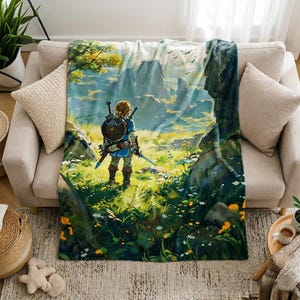 May include: A soft fleece throw blanket featuring a vibrant, full-color illustration of a character with blonde hair standing in a grassy field, holding a sword and shield, looking out toward distant mountains under a bright, clear sky.