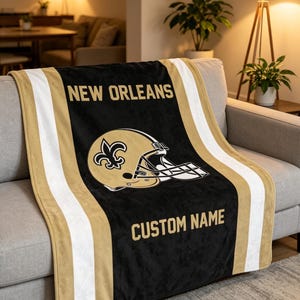 May include: A black, gold, and white fleece throw blanket featuring the text NEW ORLEANS at the top, a gold football helmet with a fleur-de-lis logo in the center, and the text CUSTOM NAME at the bottom, bordered by vertical gold and white stripes.