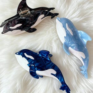 Op de afbeelding: Drie orka-vormige haarklemmen in zwart, donkerblauw en lichtblauw. Elke klem heeft een glanzende afwerking met witte markeringen op de buik en zijkanten, en een zichtbaar metalen veermechanisme voor het vastzetten van het haar.