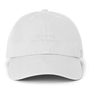 May include: A solid white baseball cap featuring a structured six-panel design with a curved brim and a button on top. The front of the cap displays the text SEO is my love language embroidered in a subtle white thread that matches the fabric.