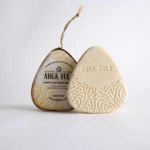 May include: Two triangular bars of off-white soap. One bar is wrapped in paper with yellow floral patterns and the text ANGA TULA, while the other is unwrapped, featuring the embossed text ANGA TULA and a raised, intricate, maze-like pattern.