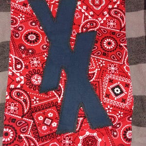 May include: A rectangular fabric patch featuring a vibrant red bandana print with white and black paisley patterns. Centered on the fabric is a large, dark blue letter X appliquéd with a visible, dark-colored zigzag stitch along the edges.