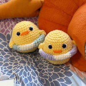 May include: Two small, round, yellow crocheted ducklings with black plastic eyes and orange stitched beaks. Each duckling wears a small, multi-colored crocheted ring around its middle, one in shades of blue and white, the other in purple and white.
