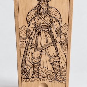 Puede incluir: Una caja de madera clara con tapa deslizante que presenta una ilustración grabada con láser de un guerrero vikingo con casco de cuernos, capa con borde de piel y túnica, sosteniendo un hacha. El nombre RAGNAR está grabado en la parte inferior.