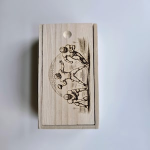 Puede incluir: Una caja rectangular de madera clara con tapa deslizante que presenta una ilustración grabada con láser en color marrón oscuro de tres jugadores de fútbol americano en acción, incluyendo un jugador central con un balón y un gráfico grande.