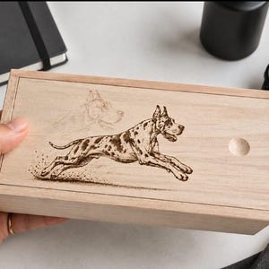 Puede incluir: Una caja rectangular de madera clara con tapa deslizante que presenta una ilustración grabada a láser en color marrón oscuro de un perro gran danés moteado en posición de carrera. La tapa incluye una pequeña muesca circular.