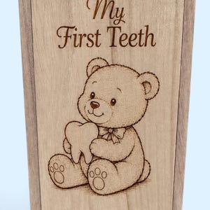 Puede incluir: Una caja rectangular de madera de color marrón claro con tapa deslizante. La tapa presenta una ilustración grabada con láser de un oso de peluche sosteniendo un diente, con el texto "My First Teeth" grabado encima. Hay una pequeña muesca circular.