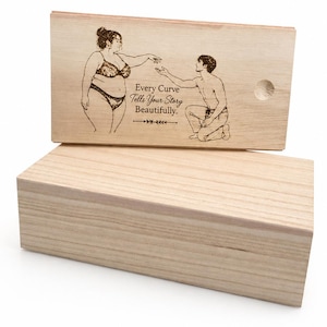 May include: A rectangular light-coloured wooden box with a sliding lid. The lid features a laser-engraved illustration of a person in lingerie standing and a person kneeling, with the text "Every Curve Tells Your Story Beautifully."
