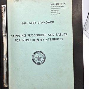 May include: A light blue document titled MIL-STD-105A, 11 September 1950, Sampling Procedures and Tables for Inspection by Attributes, featuring the Department of Defense seal, secured within a black binder with a metal spine mechanism.