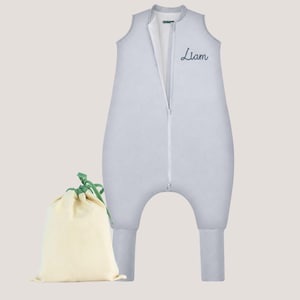 May include: A light gray sleeveless sleep sack with integrated leg openings and a central white zipper. The name Liam is embroidered in dark blue script on the upper chest. A small, cream-colored drawstring storage bag with green ties is included.