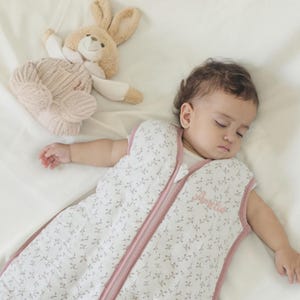 Könnte beinhalten: Ein weißer ärmelloser Schlafsack mit einem zarten braunen Blumenmuster und einem rosa Reißverschluss in der Mitte. Der Name Amelia ist in rosa Schreibschrift auf der Brust eingestickt. Ein beiges Plüschkaninchen liegt in der oberen Ecke.