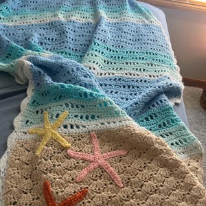 May include: A handmade crochet blanket with a beach theme. The blanket features a gradient of blue and white, resembling the ocean, and a beige section with three starfish appliqués in yellow, pink, and orange. The blanket is made of soft yarn.