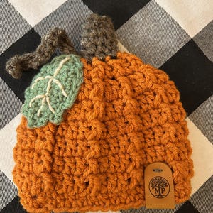 May include: Handmade orange crochet pumpkin hat with a green leaf and brown stem. The hat has a textured design and a small leather tag with a tree logo. Perfect for fall and Halloween.