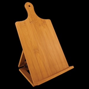 May include: A light brown bamboo wood cookbook stand shaped like a cutting board with a handle at the top, featuring a hinged support leg on the back and a horizontal ledge at the bottom to hold books or tablets in an upright, angled position.