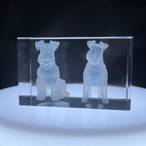 May include: A rectangular clear glass block featuring a three-dimensional laser-etched design of two terrier dogs. One dog is depicted in a seated position on the left, while the second dog stands on the right, both rendered in frosted white.