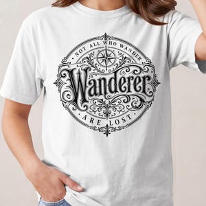 以下が含まれることがあります： 胸元に黒い円形のグラフィックがプリントされた白い半袖Tシャツ。デザインには装飾的なスクロール模様、上部にコンパスローズ、中央の「Wanderer」という文字を囲むように「NOT ALL WHO WANDER ARE LOST」というテキストが配置されています。