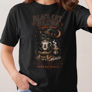 May include: A black t-shirt featuring a graphic of a black cat wearing a witch hat, sitting on a stool and holding a coffee mug. The design includes a cauldron, a crescent moon, and the text "BLACK CAT BREWS COFFEE HOUSE" and "HEX-PRESSO SERVED DAILY."