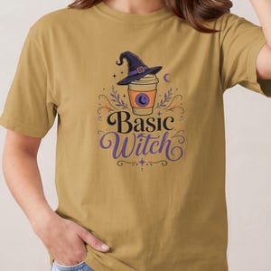 May include: A mustard yellow short-sleeve t-shirt featuring a graphic of a coffee cup wearing a purple witch hat. The text "Basic Witch" is printed below the cup in a black and purple font, surrounded by decorative leaves, stars, and moons.