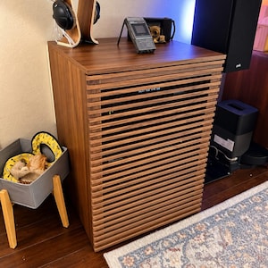 May include: A tall, rectangular wooden media cabinet with horizontal slats covering the front. On top sits a silver digital voice recorder and a wooden headphone stand with black over-ear headphones. A gray fabric bin with wooden legs is nearby.
