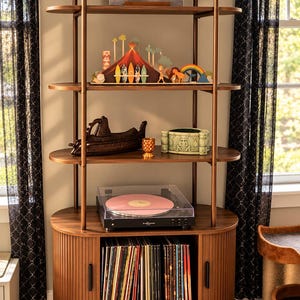 May include: A tall, brown wooden shelving unit with arched top supports, featuring four shelves and a lower cabinet with tambour doors. The unit holds a vinyl record player with a pink disc, a collection of records, and various decorative items.