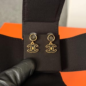 May include: A pair of gold-tone drop earrings featuring a circular stud with a small star detail and a dangling interlocking CC logo. The design incorporates black enamel accents on the logo and the circular stud, set against a black fabric display.
