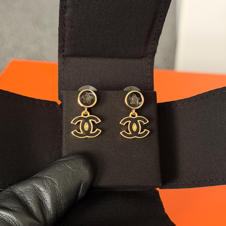 May include: A pair of gold-tone drop earrings featuring a circular stud with a small star detail and a dangling interlocking CC logo. The design incorporates black enamel accents on the logo and the circular stud, set against a black fabric display.