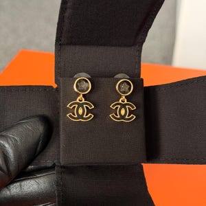 May include: A pair of gold-tone earrings featuring a circular stud with a small star cutout, connected to a dangling interlocking CC logo charm. The earrings are finished with black enamel detailing and are presented on a black fabric display card.