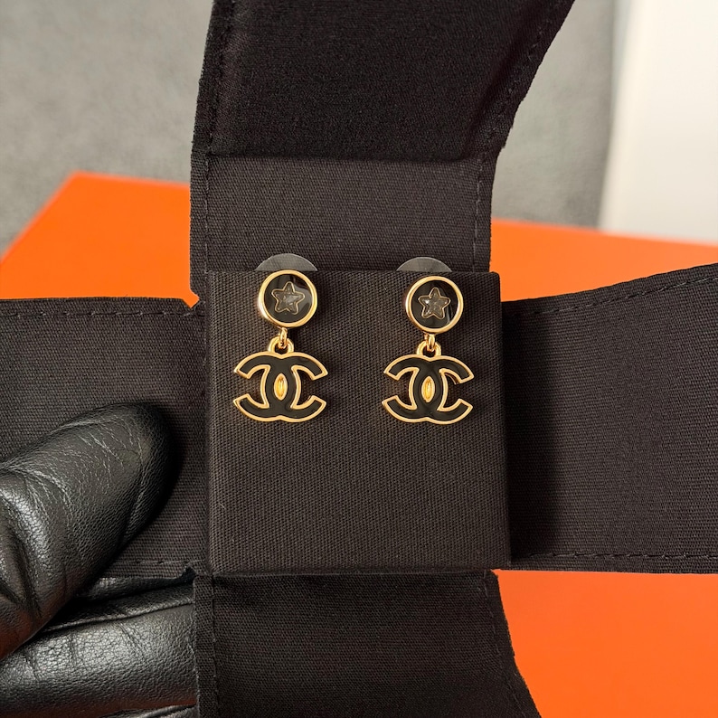 May include: A pair of gold-tone earrings featuring a circular stud with a small star cutout, connected to a dangling interlocking CC logo charm. The earrings are finished with black enamel detailing and are presented on a black fabric display card.