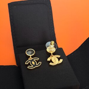 May include: A pair of gold-tone earrings featuring a circular stud with a black star inlay and a dangling interlocking CC logo charm. The earrings are displayed on a black velvet-textured insert, showcasing a polished gold finish and black enamel details.
