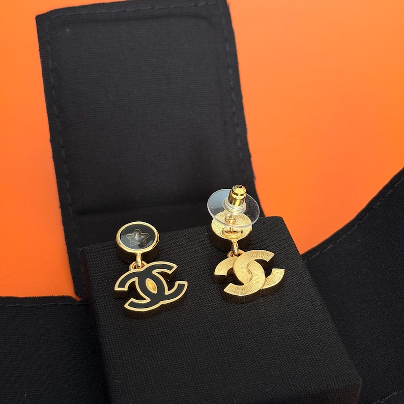 May include: A pair of gold-tone earrings featuring a circular stud with a black star inlay and a dangling interlocking CC logo charm. The earrings are displayed on a black velvet-textured insert, showcasing a polished gold finish and black enamel details.