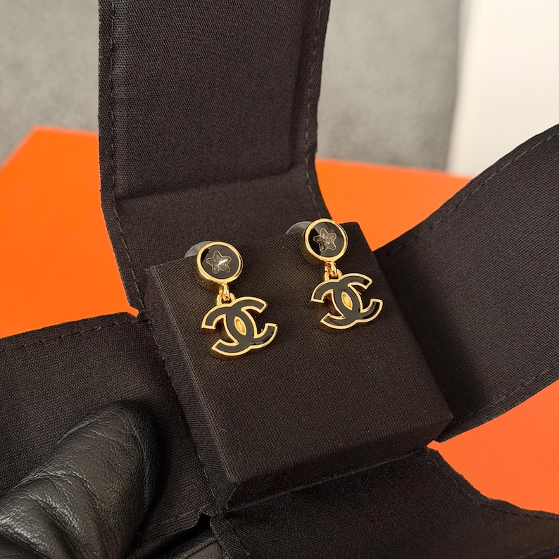 May include: A pair of gold-tone drop earrings featuring a circular stud with a small star cutout and a dangling interlocking CC logo. The earrings are finished with black enamel detailing on both the circular stud and the logo charm, presented in a box.