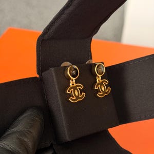 May include: A pair of gold-tone drop earrings featuring a circular stud with a black inlay and a small star detail. A dangling gold-tone interlocking CC logo charm is attached to each stud. The earrings are presented on a black fabric display card.