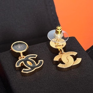 May include: A pair of gold-tone drop earrings featuring a circular stud with a black inlay and a small star detail, connected to a dangling interlocking CC logo charm. One earring shows a black enamel finish on the logo, while the other displays a textured gold-tone metal surface. Each earring includes a clear plastic disc backing.