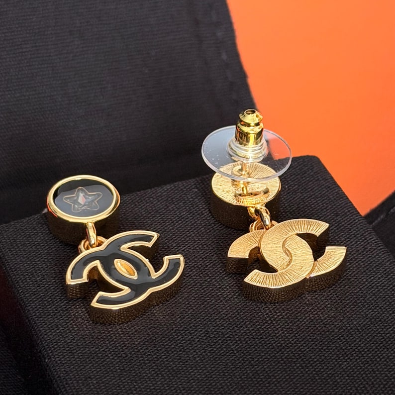 May include: A pair of gold-tone drop earrings featuring a circular stud with a black inlay and a small star detail, connected to a dangling interlocking CC logo charm. One earring shows a black enamel finish on the logo, while the other displays a textured gold-tone metal surface. Each earring includes a clear plastic disc backing.