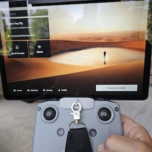 Op de afbeelding: Een grijze drone-afstandsbediening met een zwarte draagriem bevestigd via een zilveren metalen clip. Een tablet is bovenop de controller gemonteerd en toont een digitale interface met de tekst Before You Fly, QuickTransfer en Connection Guide.