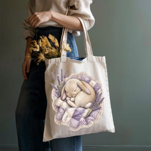May include: A cream-colored canvas tote bag featuring a printed illustration of a light brown rabbit sleeping on a purple and white patchwork quilt, surrounded by sprigs of lavender flowers, with two long shoulder straps for carrying.