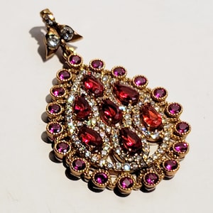 May include: A gold pendant with a teardrop shape, featuring a cluster of red and clear gemstones. The pendant is surrounded by a border of smaller pink gemstones.