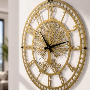 Può includere: Un orologio da parete circolare con una cornice in metallo dorato caratterizzata da un intricato design ritagliato di un albero al centro. L'orologio ha lancette nere per ore, minuti e secondi e grandi numeri arabi ritagliati da uno a dodici.
