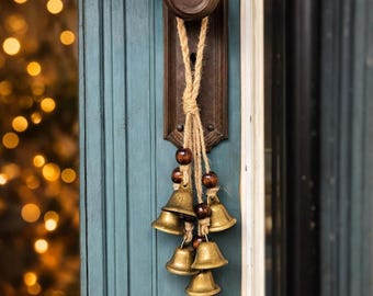 May include: A hanging decoration featuring multiple brass-colored metal bells suspended by natural jute twine. The arrangement includes several dark brown wooden beads interspersed along the twine, creating a cluster of bells that hang vertically.