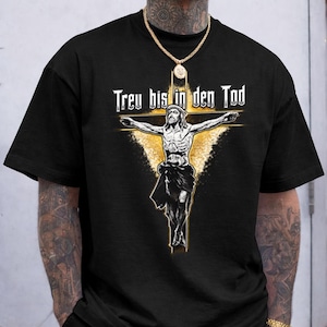 Puede incluir: Una camiseta negra de manga corta con un estampado gráfico de una figura en una cruz con un efecto de luz dorada detrás. El texto "Treu bis in den Tod" está impreso en blanco sobre la figura. La camiseta tiene un cuello redondo estándar.