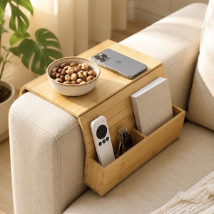 May include: A light-coloured bamboo sofa armrest tray with a flat top surface and a side storage compartment. The tray holds a ceramic bowl filled with nuts, a smartphone, a white remote control, a set of keys, and a small notebook.