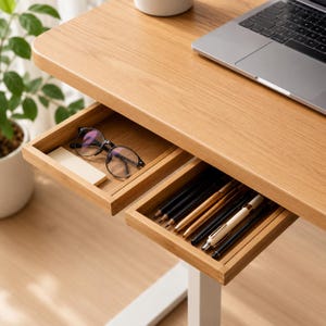 Op de afbeelding: Een lichthouten bureau met twee open lades. De linkerlade bevat een bril met een zwart montuur en een klein rechthoekig briefje. De rechterlade bevat een verzameling diverse pennen en potloden. Een laptop staat op het bureauoppervlak.